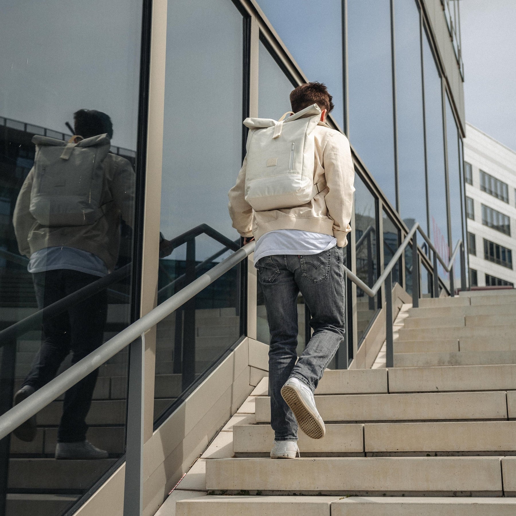 Mann trägt weißen Rolltop-Rucksack 'Robin Medium' und steigt Außenstiege neben Glasfassade hoch
