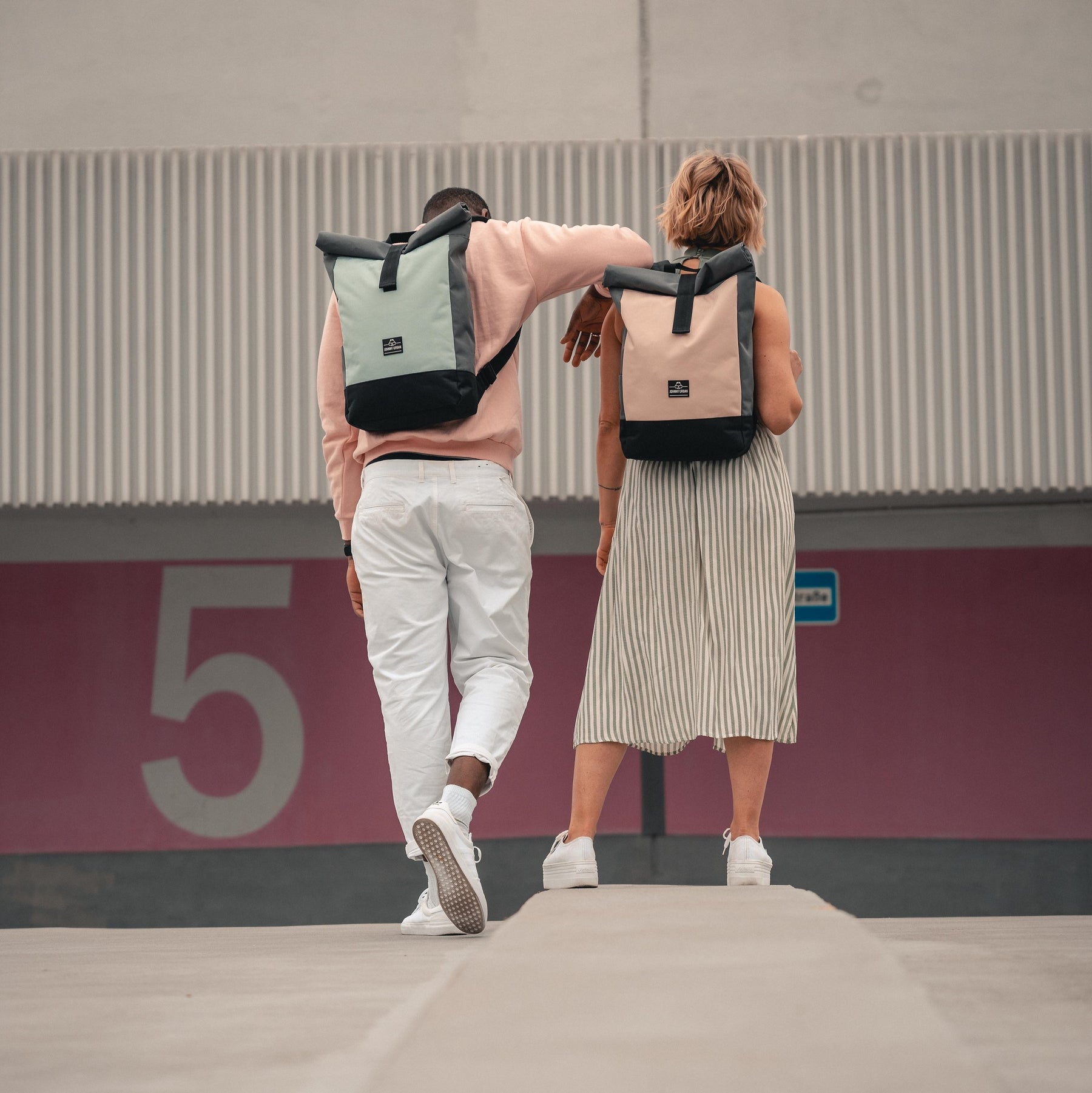 Zwei Personen von hinten mit Rolltop Rucksack 'Ryan' in Pastellfarben, stehend vor einer Wand mit großer Zahl 5