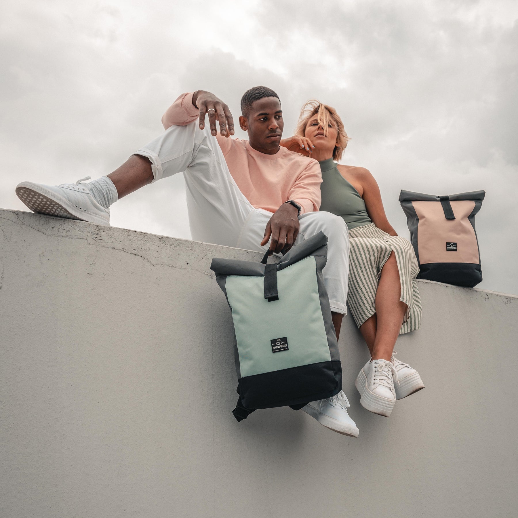Junger Mann und Frau sitzen auf einer Mauer mit Rolltop Rucksäcken 'Ryan' in Pastell-Mint und Pastell-Rosa