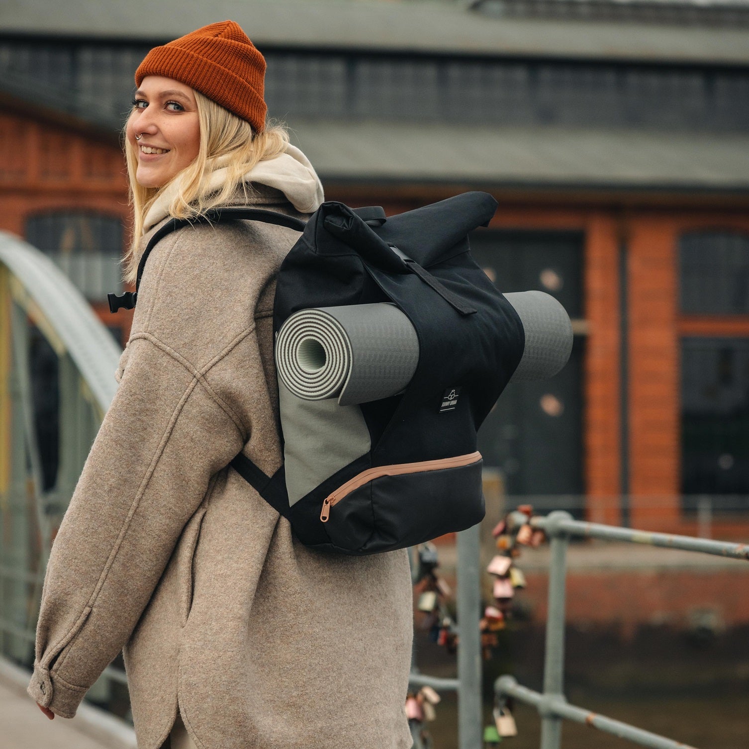 Frau mit oranger Mütze und beige Jacke trägt schwarzen Rolltop Rucksack 'Allen Large' mit grauer Yogamatte an urbanem Geländer
