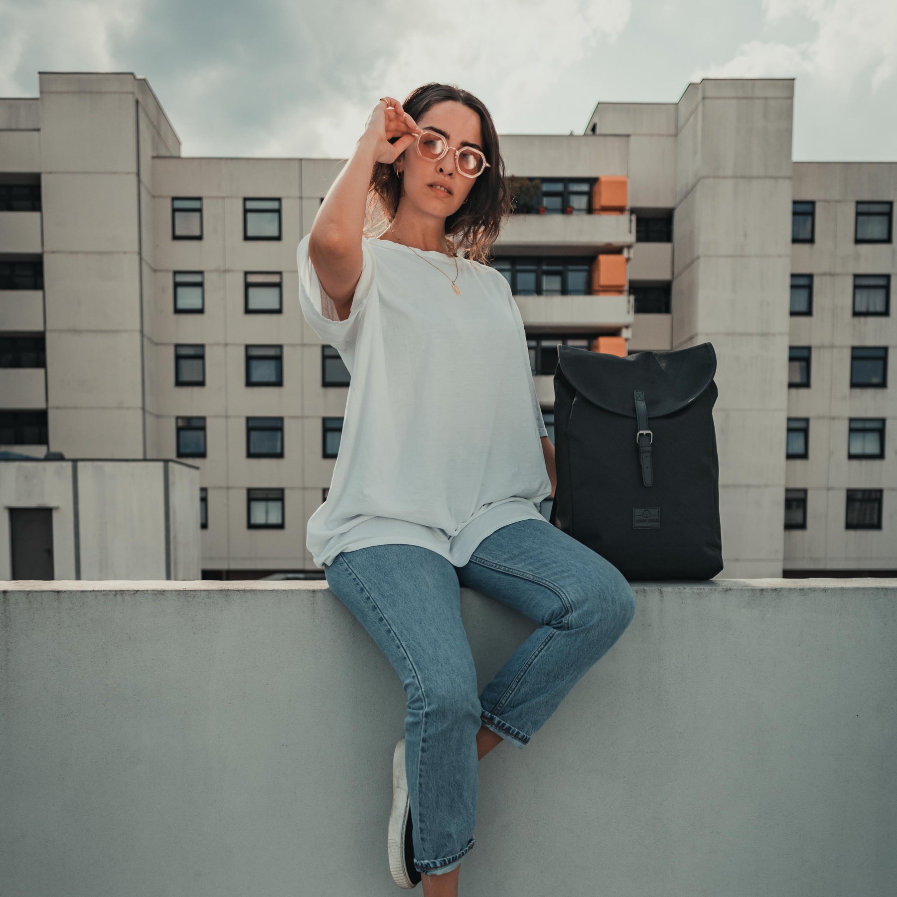 Frau mit weißem T-Shirt und blauen Jeans sitzt auf einer Mauer neben schwarzem Rucksack 'Liam' von Johnny Urban
