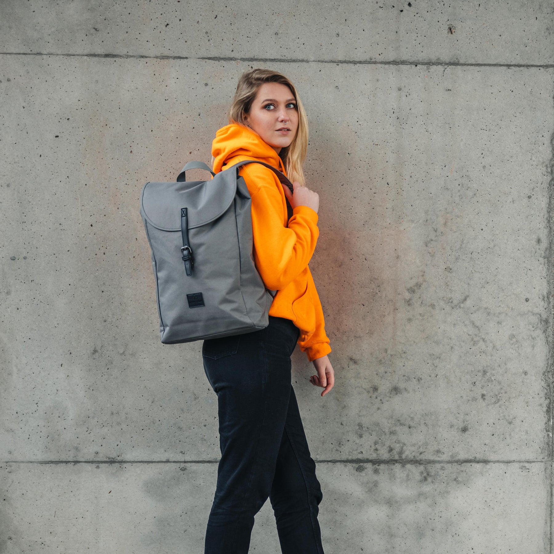 Frau mit dunkelgrauem Rucksack 'Liam' trägt orangefarbenen Hoodie vor grauer Betonwand