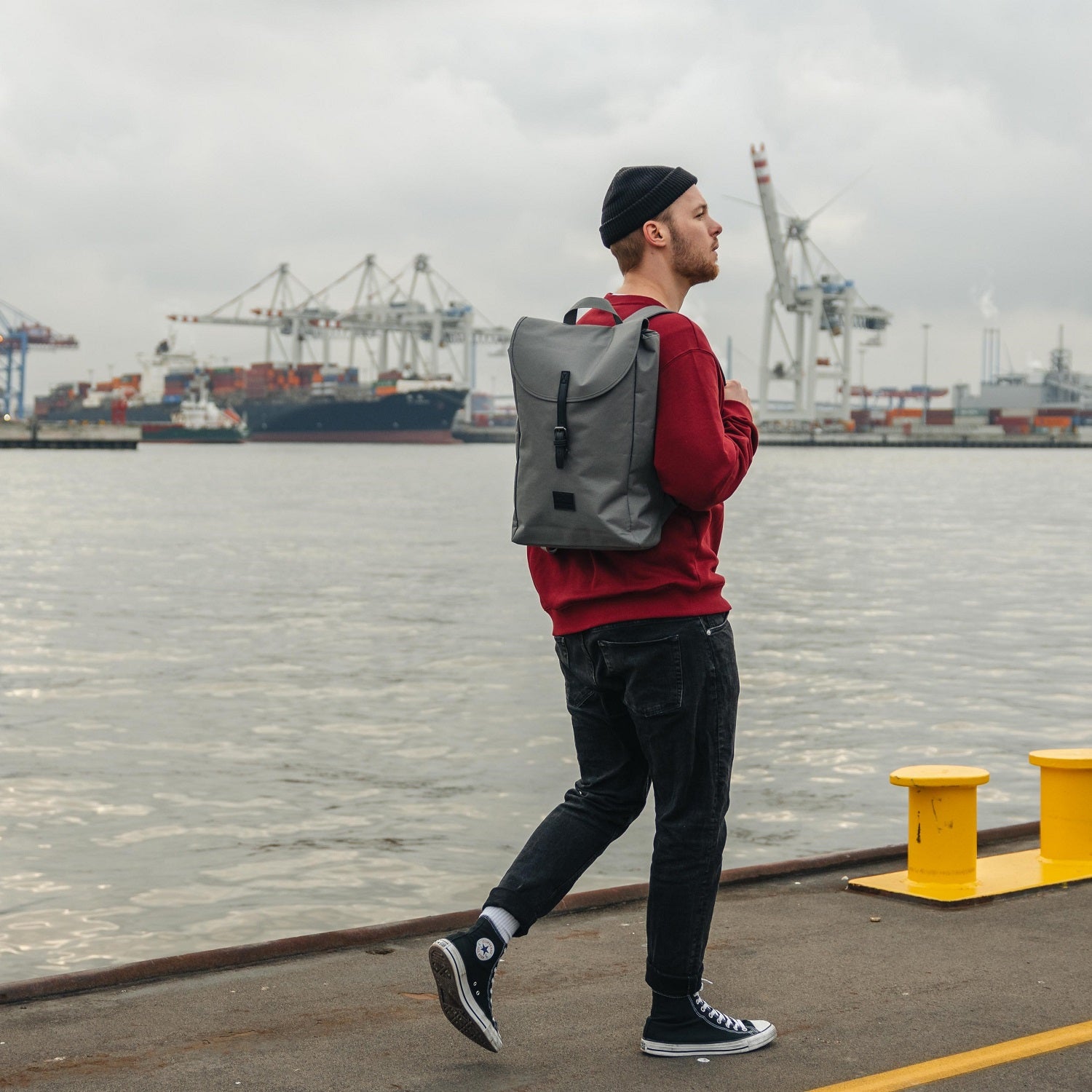 Mann am Hafen trägt dunkelgrauen Rucksack 'Liam', rote Sweatshirt, schwarze Jeans, schwarze Mütze und Converse Schuhe