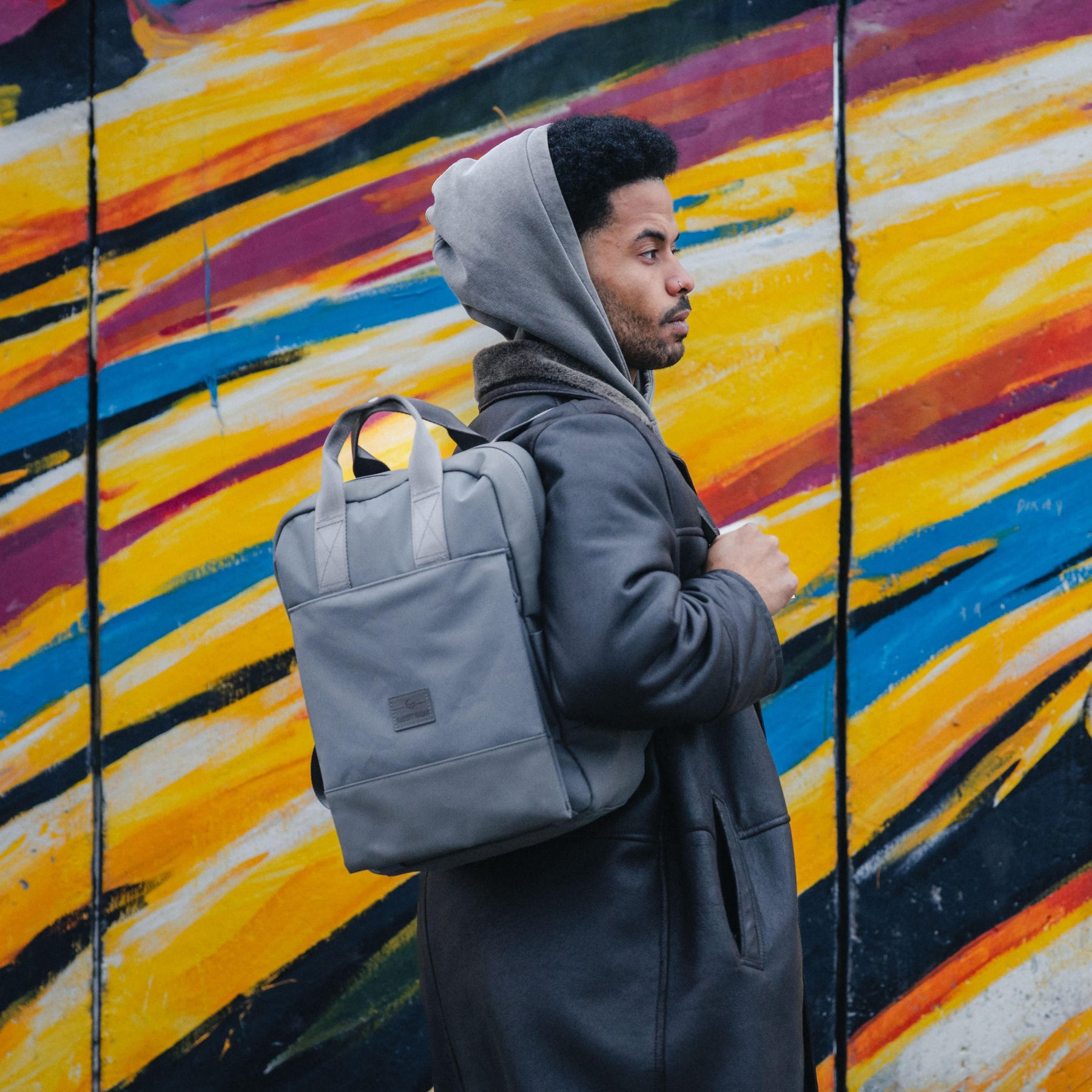 Mann mit grauem Rucksack 'Jona Large' steht vor einer bunten Wand mit gelben, blauen und roten Streifen