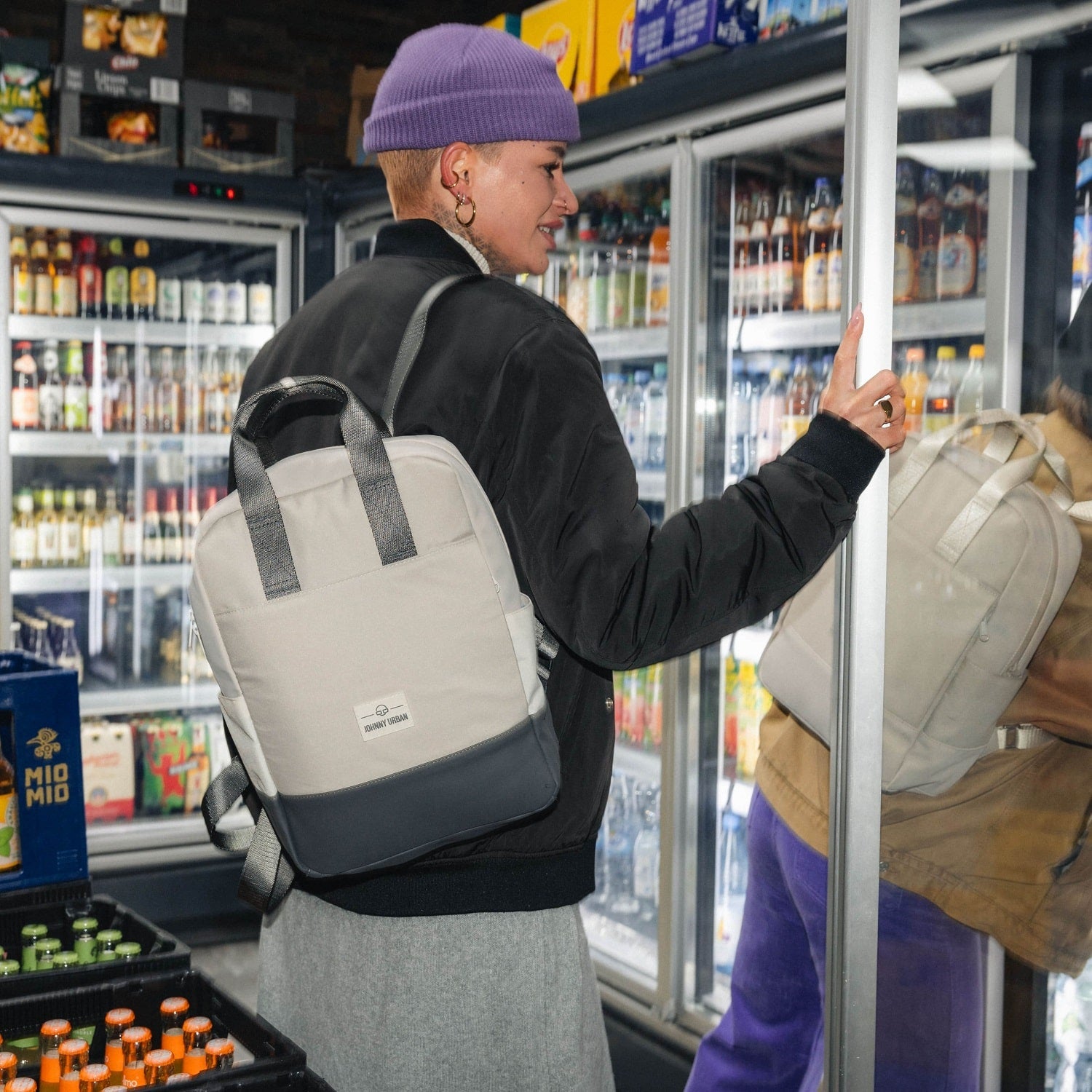 Frau mit lila Mütze und schwarzer Jacke trägt den beigen-grauen Rucksack 'Jona Medium' von Johnny Urban beim Öffnen einer Kühlschranktür