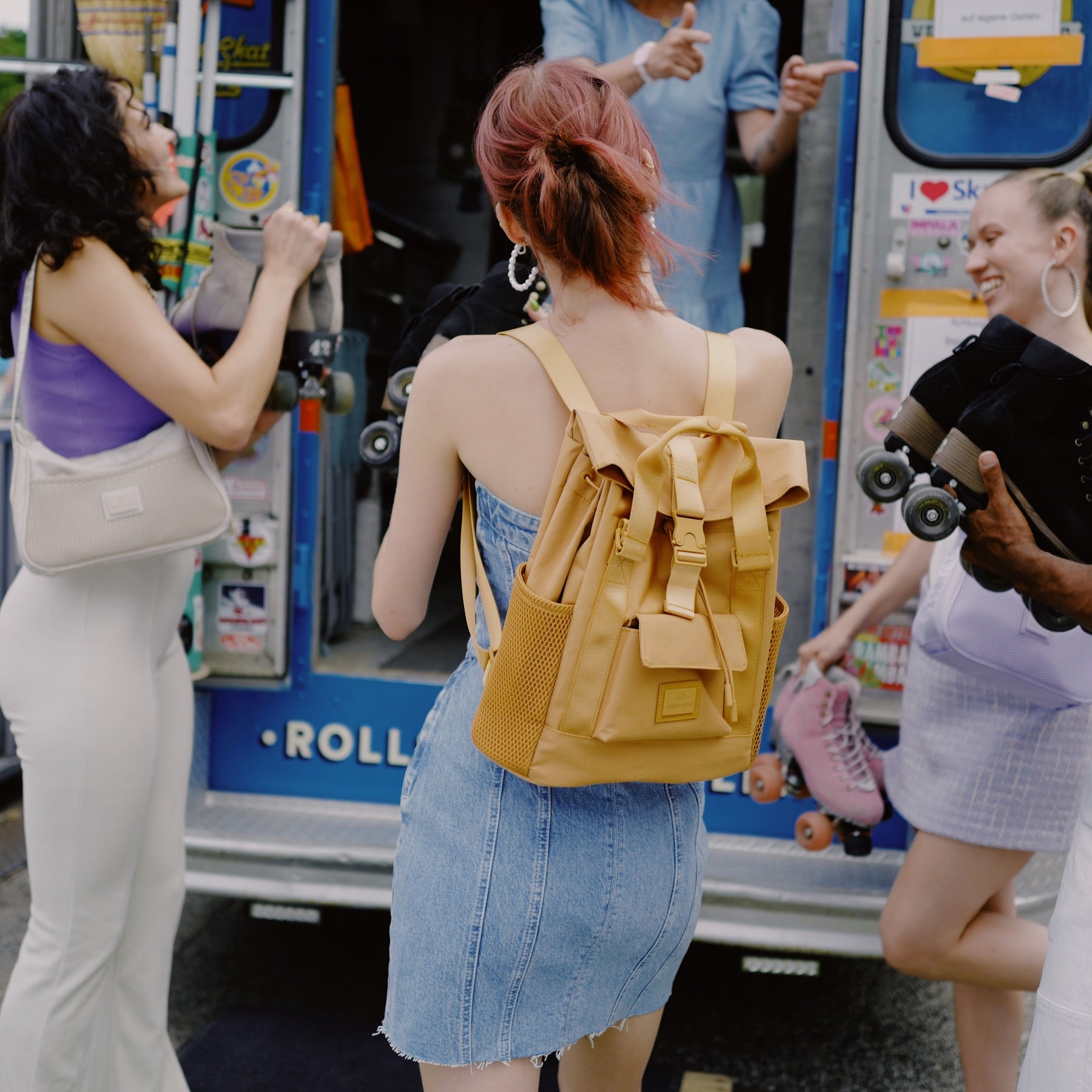 Rucksack 'Jules' in karamellbraun aus Leder auf dem Rücken einer Frau in Jeanskleid vor einem Rollschuh-Verkaufswagen