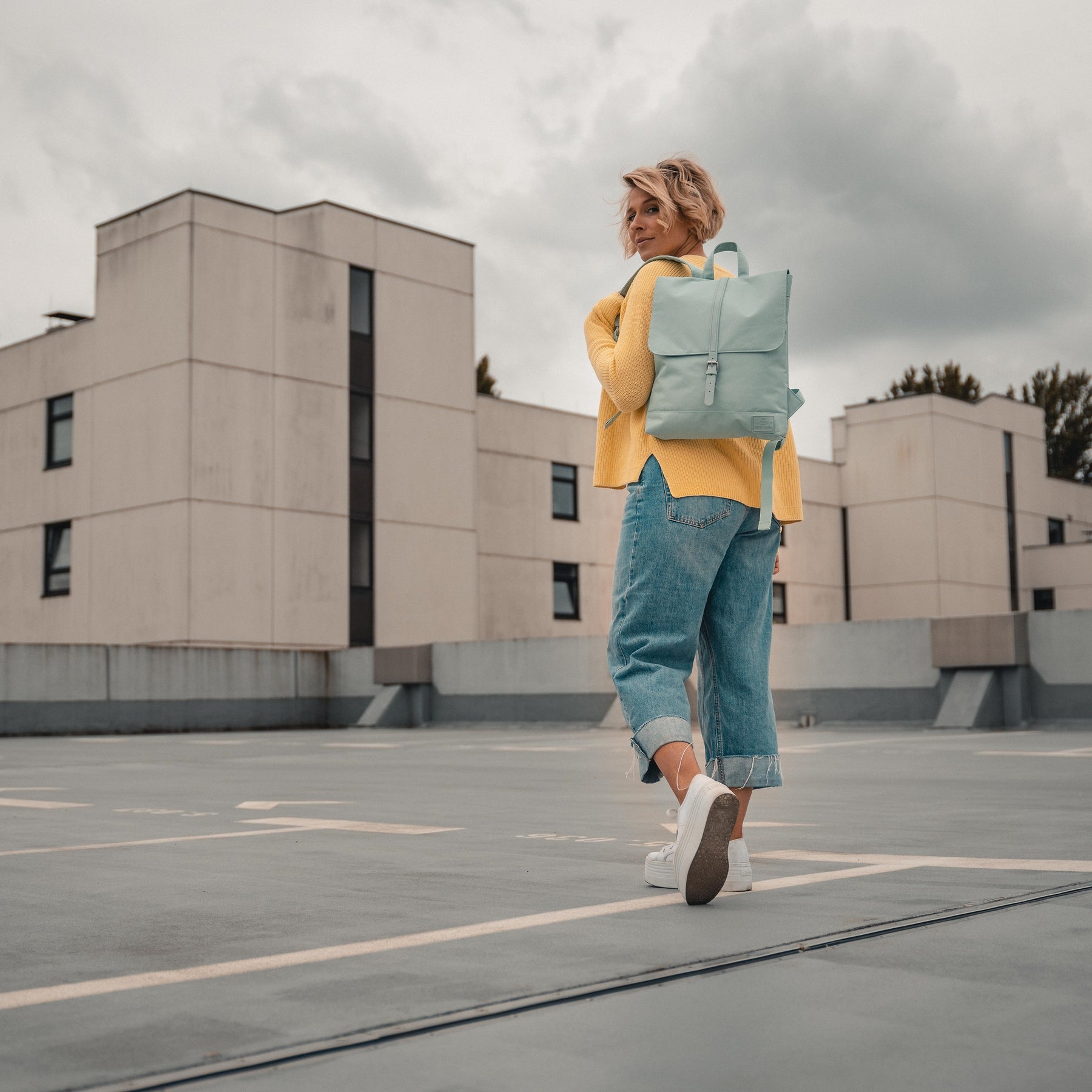 Frau trägt mintfarbigen Rucksack 'Mia', gelber Pullover, blaue Jeans, weiße Sneakers auf Dachparkplatz