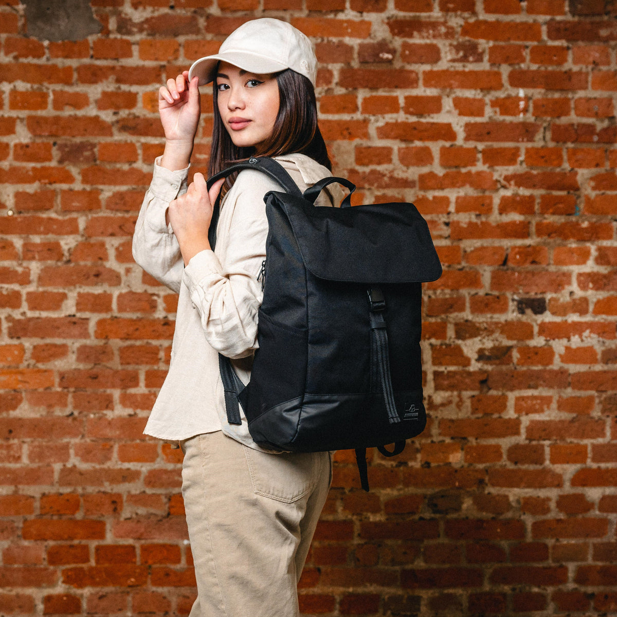 Frau mit schwarzem Rucksack 'Mika', hellbeige Cap und beiger Kleidung vor roter Ziegelwand