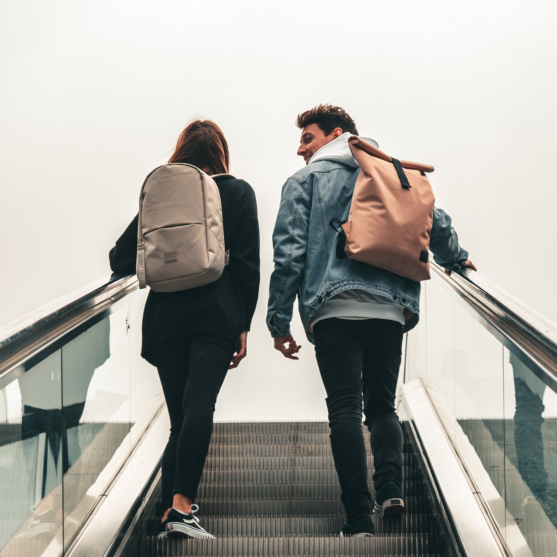 Zwei Personen auf Rolltreppe, weiblich mit beigem Rucksack 'Emil', männlich mit rosa Rolltop-Rucksack
