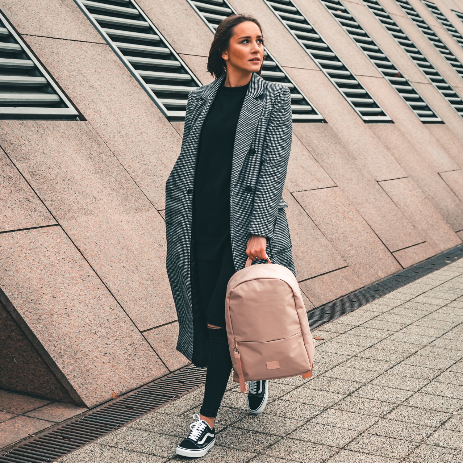Teenager-Mädchen hält rosa Rucksack 'Milo' in der Hand, trägt karierten Mantel und schwarze Sneaker