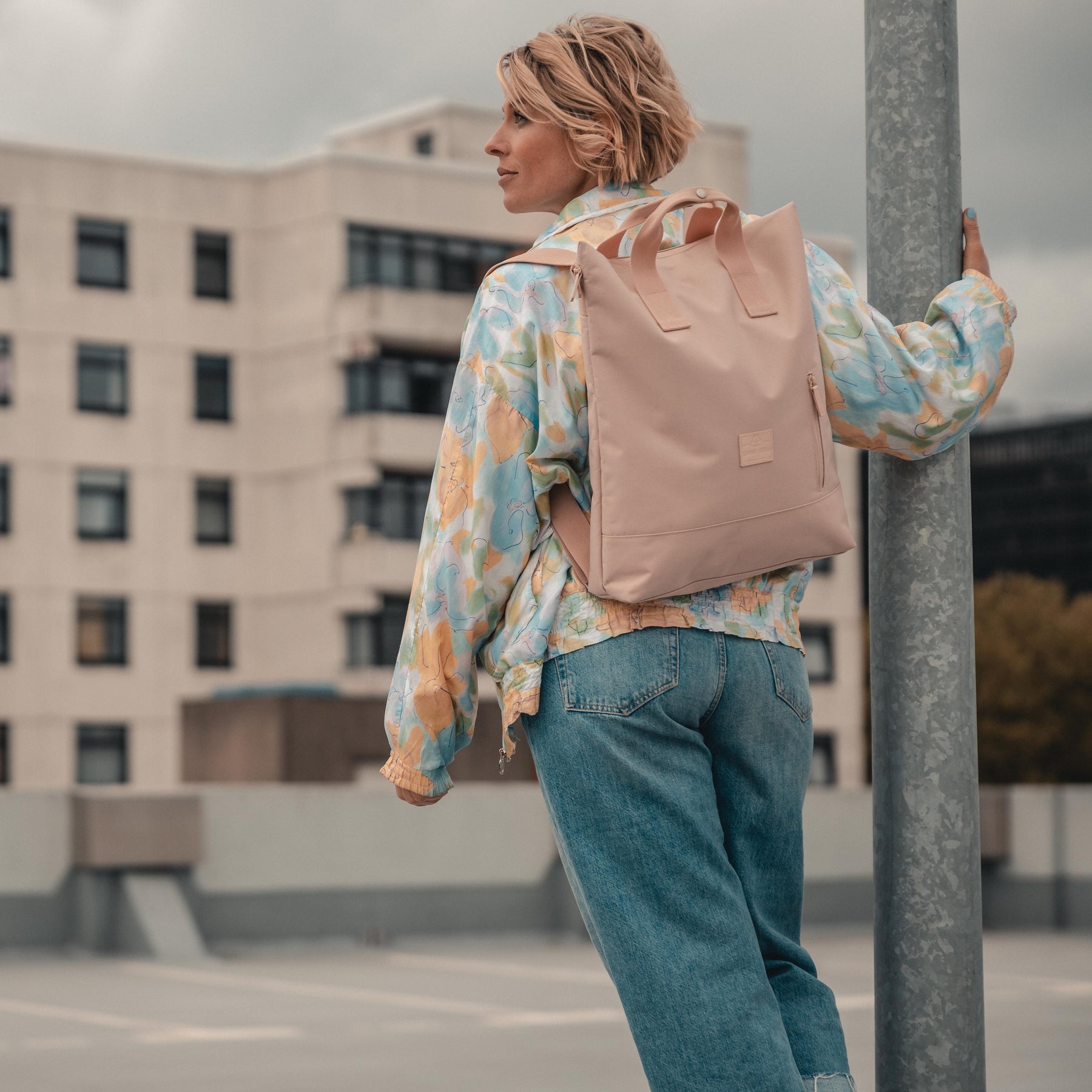 Frau trägt rosa Rucksacktasche 'Ivy' von Johnny Urban, lässige bunte Jacke und blaue Jeans, stützt sich auf Laternenpfahl