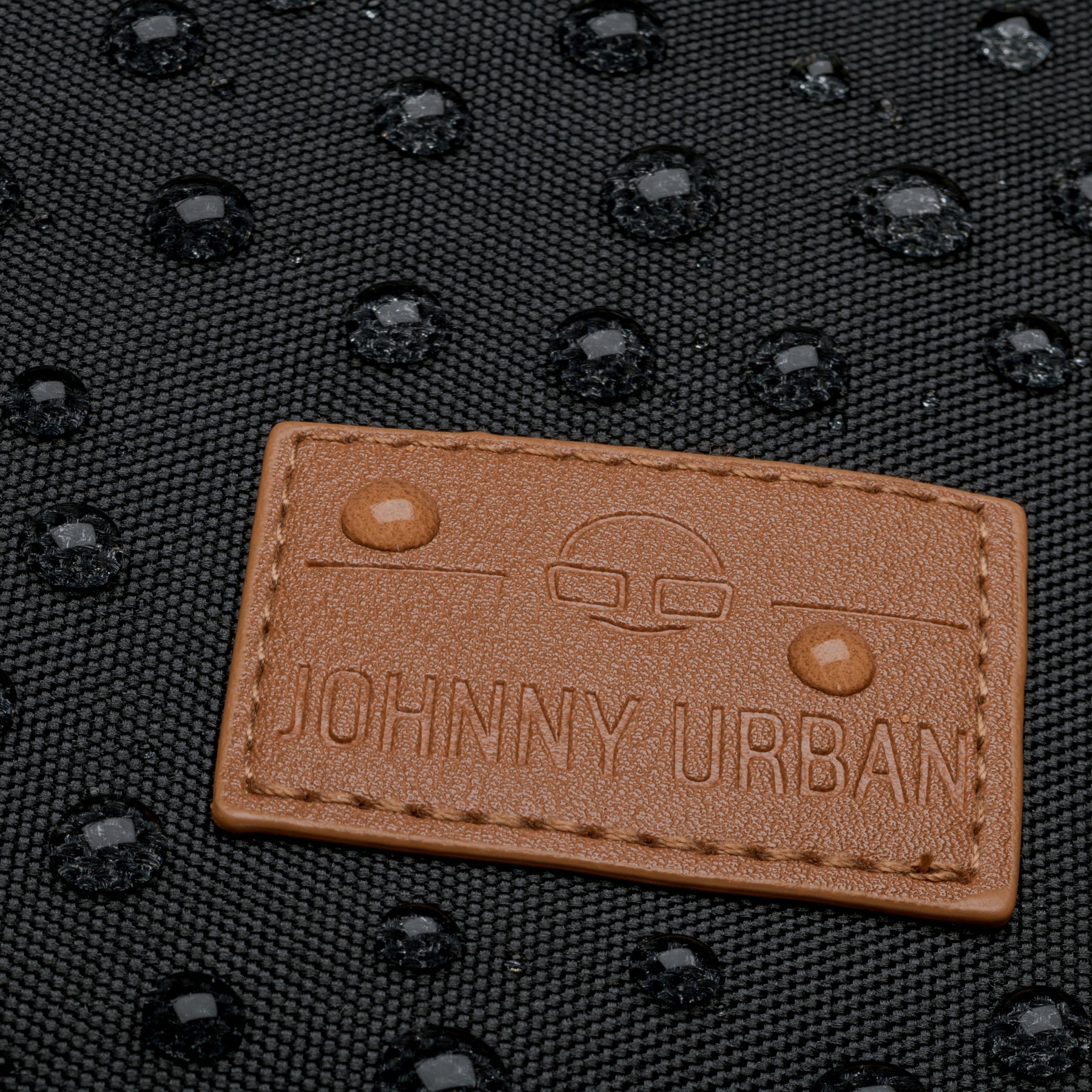 Braunes Johnny Urban Markenlabel auf wasserabweisendem schwarzen Rucksackmaterial mit Wassertropfen