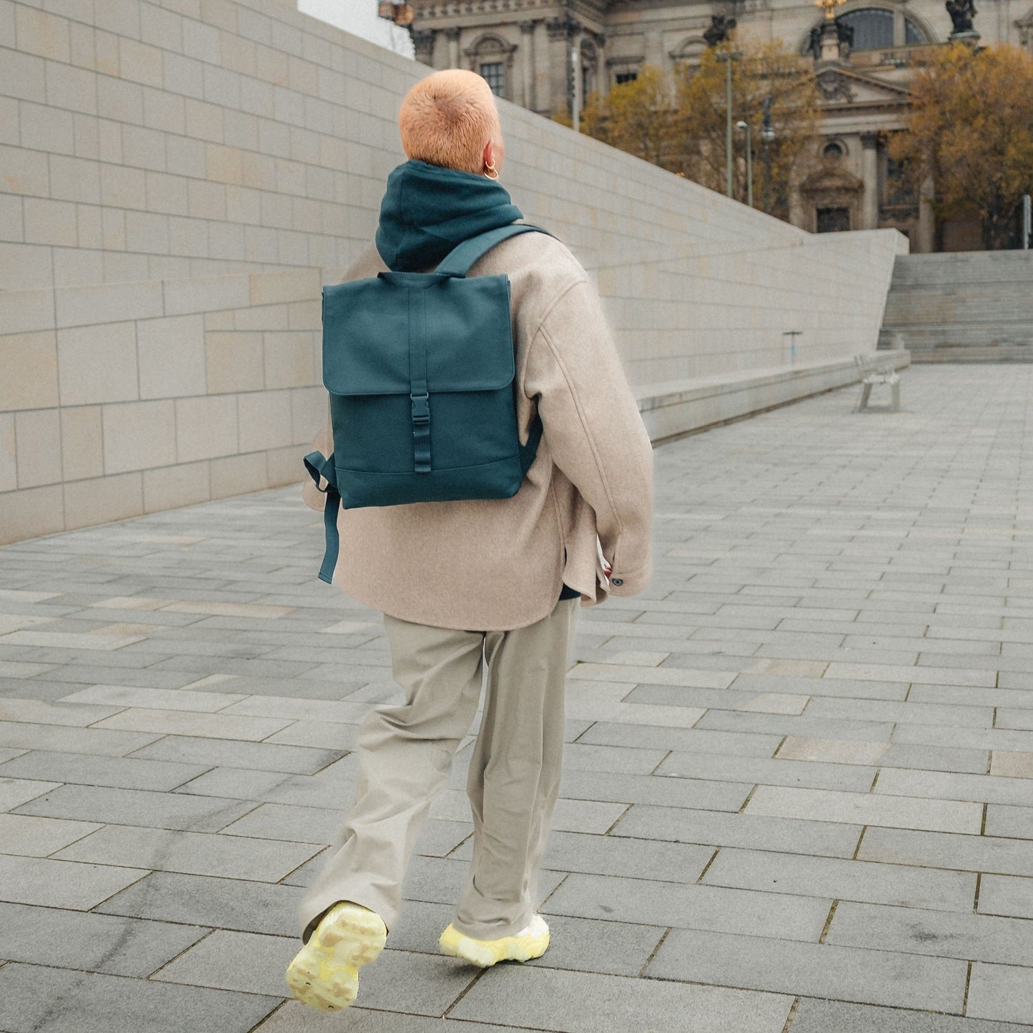 Frau mit kurzem haarschnitt trägt petrolgrünen Rucksack 'Ruby' und beige hose in urbaner umgebung