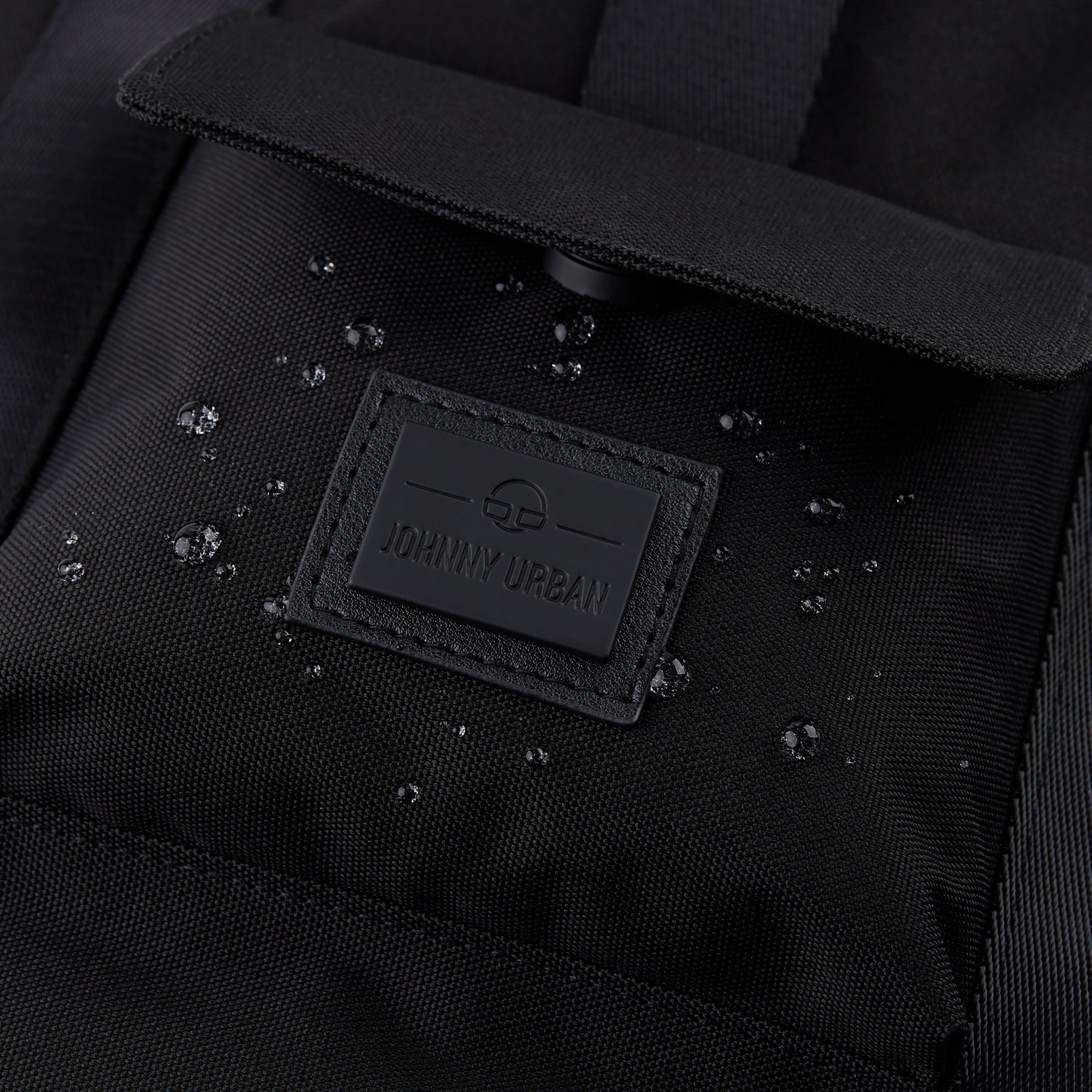 Nahaufnahme des schwarzen wasserabweisenden Rucksacks 'Jules' mit Johnny Urban Logo und Wassertröpfchen auf dem Stoff
