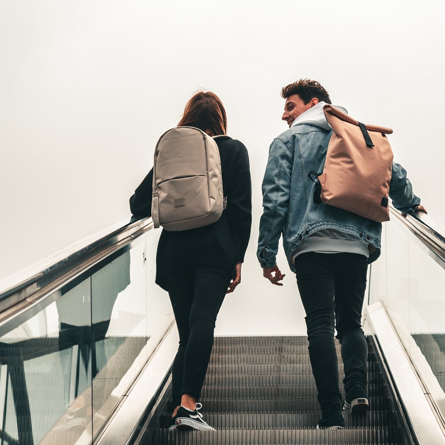 Teenager-Mädchen mit grauem Rucksack 'Milo' und junger Mann mit beigem Rucksack auf Rolltreppe