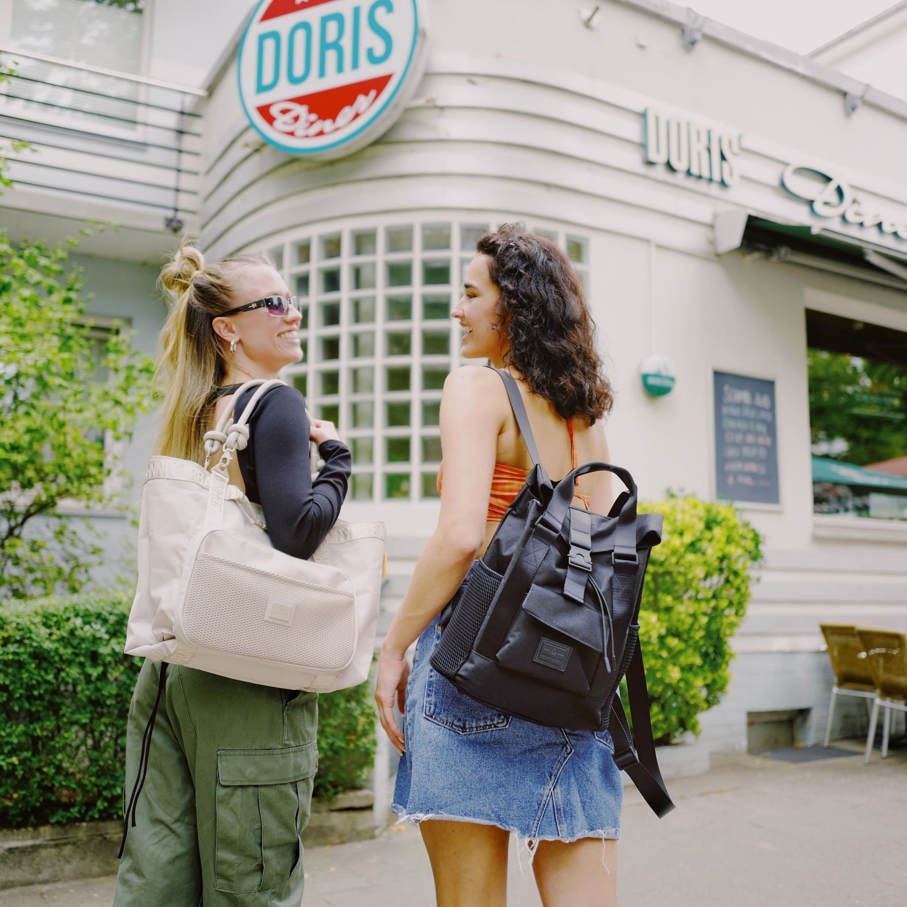 Zwei Frauen vor Café 'Doris' streiten lachend. Eine trägt beige Shopper Tasche 'Cassie', die andere schwarzen Rucksack.