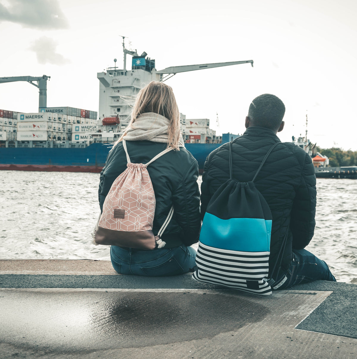 Zwei Personen sitzen mit dem Rücken zum Betrachter am Wasser, eine trägt den rosa Turnbeutel 'Noah' mit weißem geometrischem Muster
