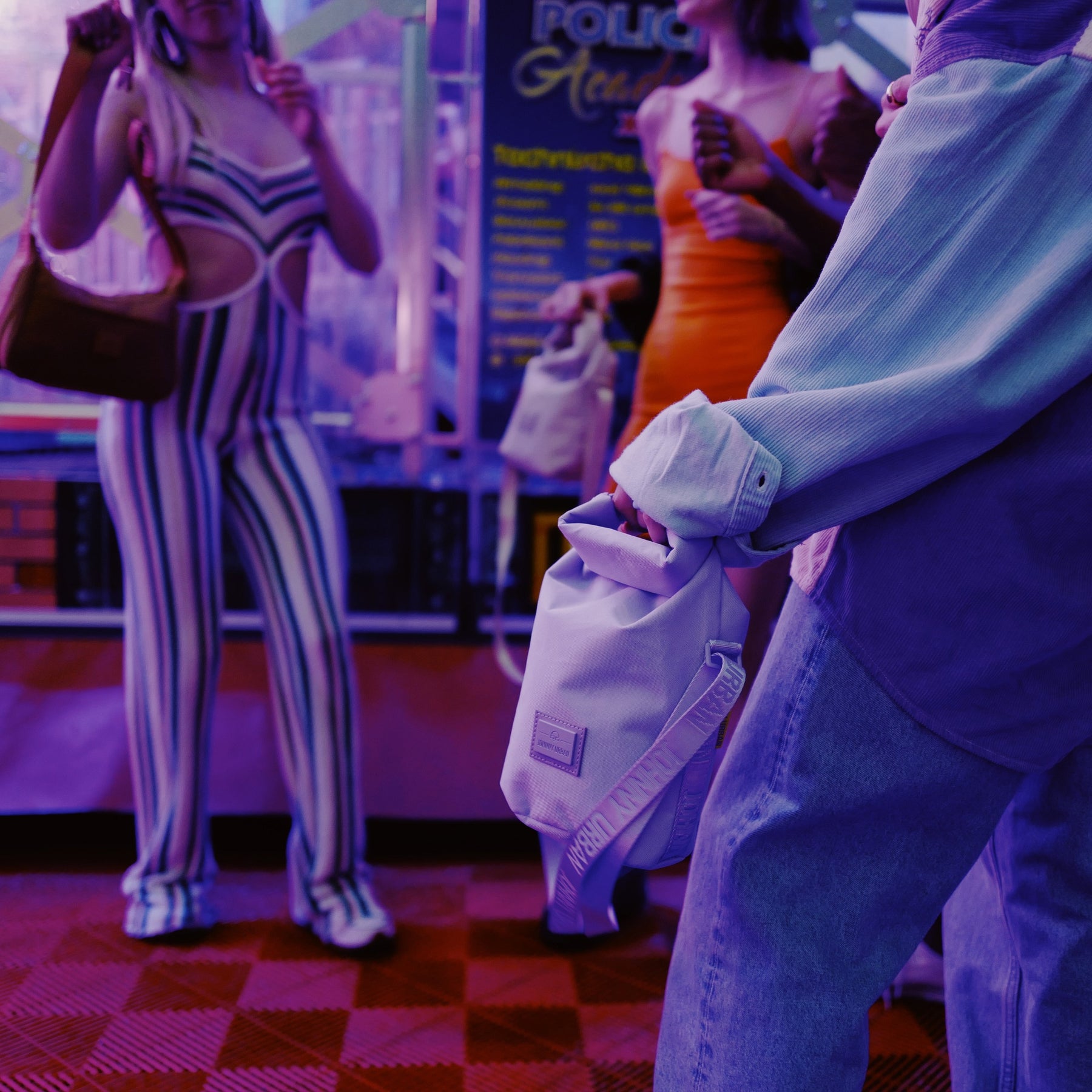 Person hält die lilafarbene Umhängetasche 'Rue' von Johnny Urban in der Hand, im Hintergrund Menschen in bunter Kleidung