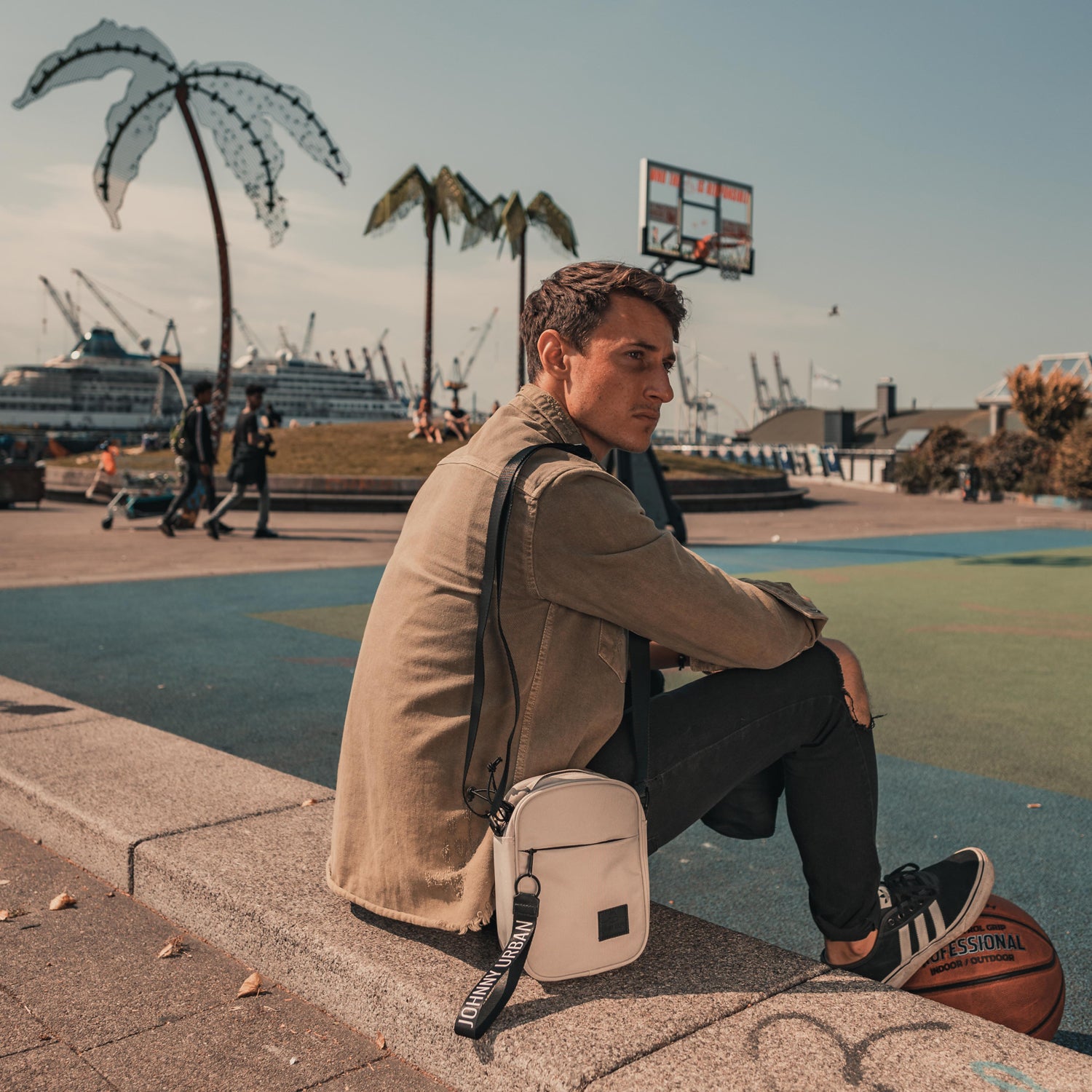 Mann sitzt an Außenbasketballfeld, trägt beige Umhängetasche 'Luis' von Johnny Urban, hält Basketball