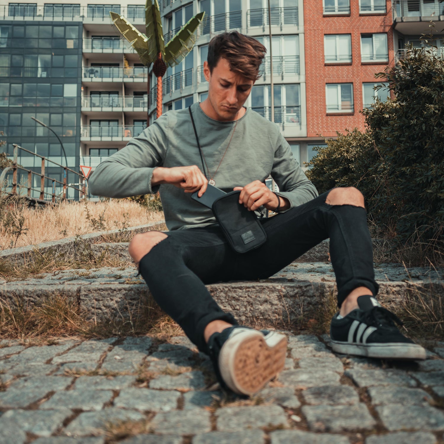 Junger Mann sitzt auf Steintreppe, trägt schwarze Umhängetasche 'Josh' und zieht etwas heraus