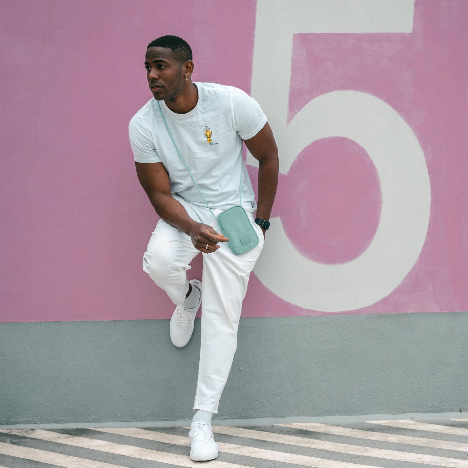 Mann trägt mintgrüne Umhängetasche 'Josh' von Johnny Urban, weiße Hose und T-Shirt vor rosa Wand mit großer Zahl 5