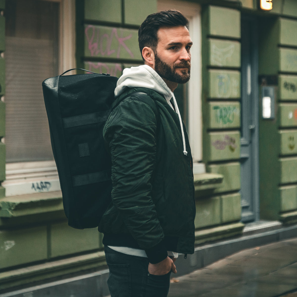 Mann mit schwarzem Weekender-Rucksack 'Paul' von Johnny Urban, grüne Jacke und grauer Hoodie in urbaner Umgebung