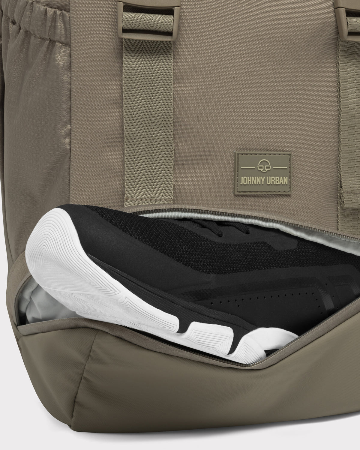 Johnny Urban Rucksack 'Nico' in desertgrau mit seitlichem Fach, das schwarze Sneaker mit weißer Sohle hält