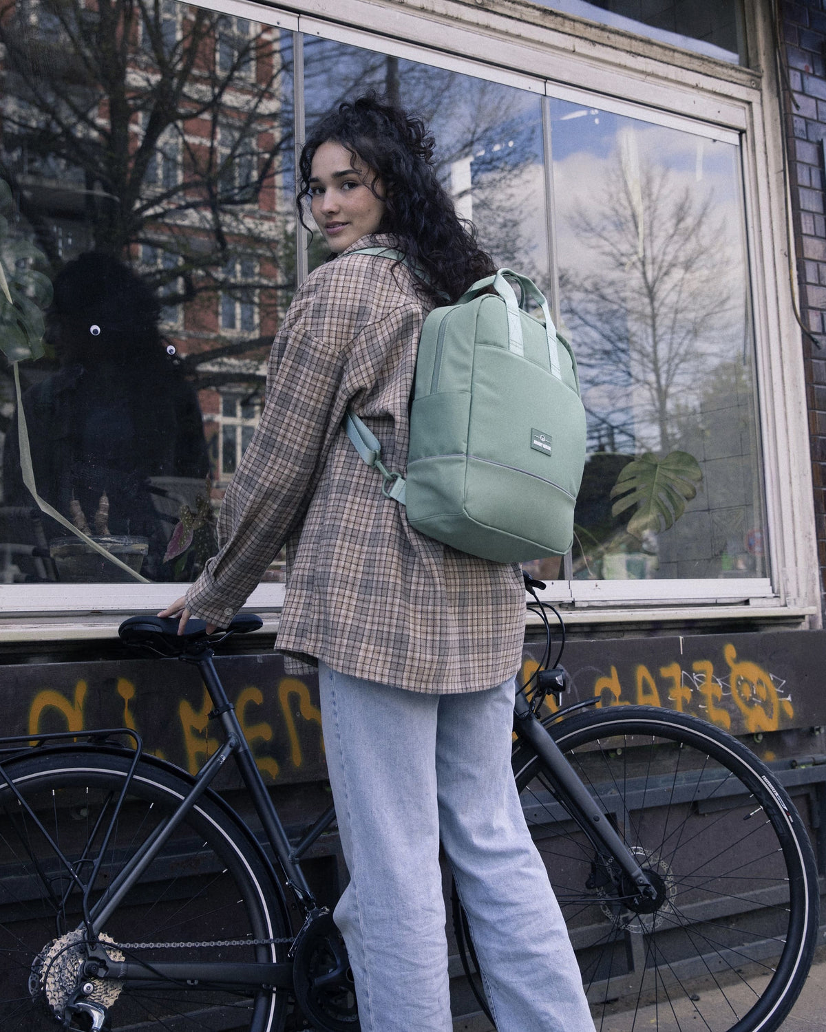 Frau mit mintgrünem Johnny Urban Rucksack Jona Medium sitzt auf Fahrrad vor Fenster mit Pflanzen