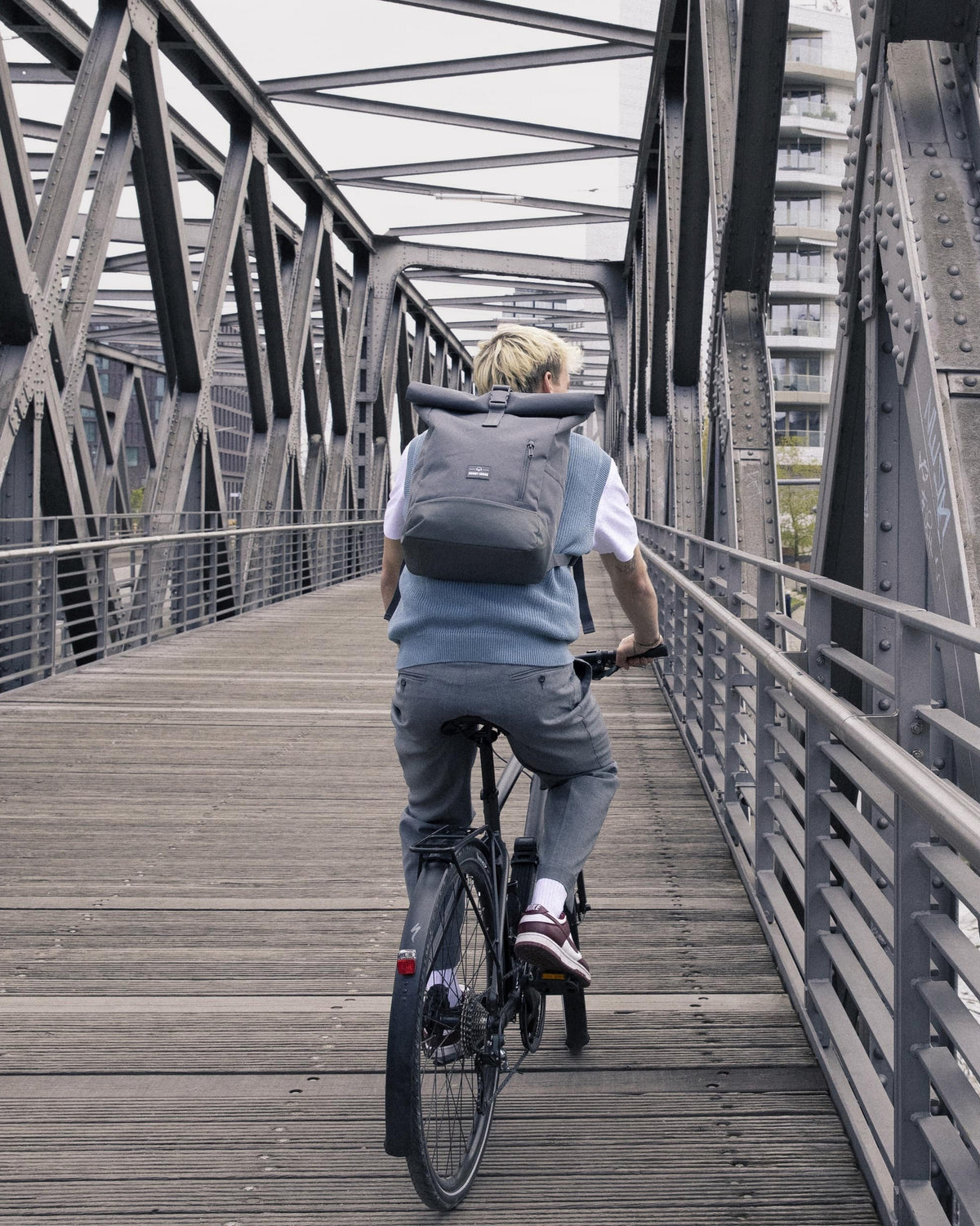Mann fährt mit Rolltop Rucksack 'Robin Medium' auf einem Fahrrad über eine stählerne Fußgängerbrücke in der Stadt