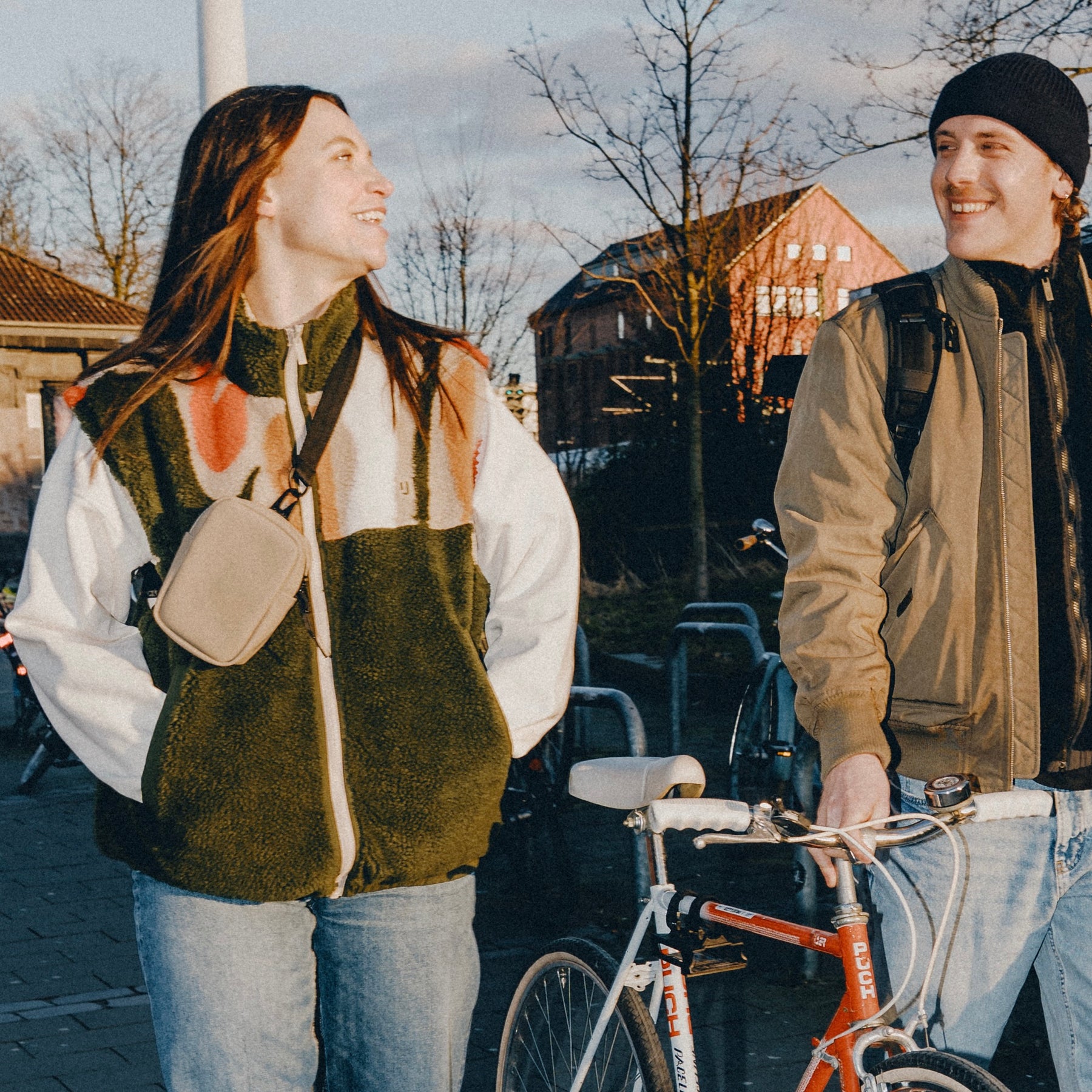 Frau mit beigem Front Pack 'Toby' von Johnny Urban trägt bunte Fleecejacke, Mann mit Fahrrad und schwarzer Mütze