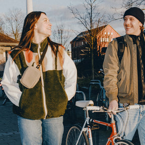Frau mit beigem Front Pack 'Toby' von Johnny Urban trägt bunte Fleecejacke, Mann mit Fahrrad und schwarzer Mütze