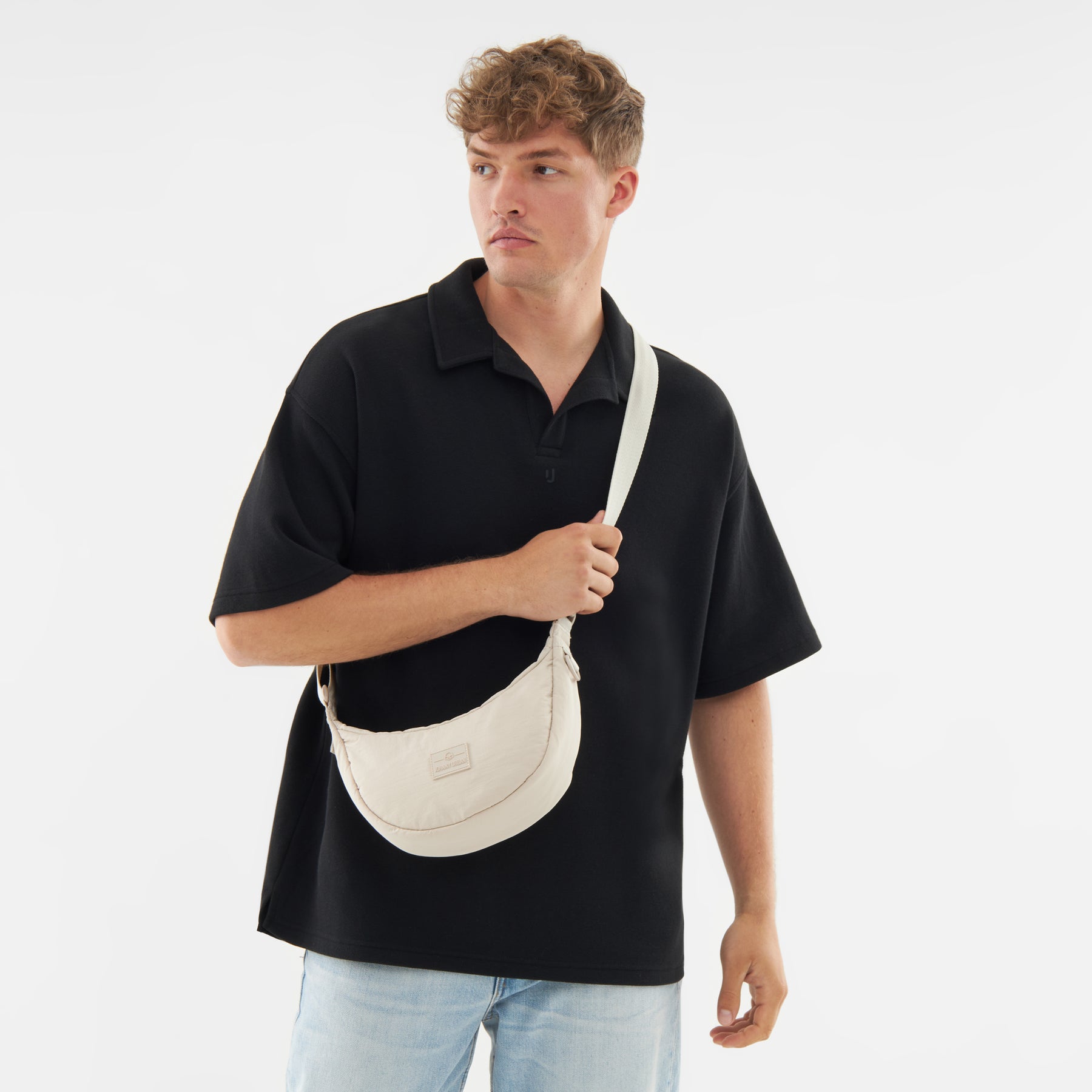 Mann trägt beige Half Moon Bag 'Kaia Small' über der Schulter, trägt schwarzen Poloshirt und helle Jeans