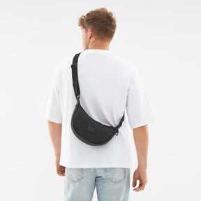 Mann trägt schwarze Half Moon Bag 'Kaia Small' quer am Rücken über weißem T-Shirt und blauer Jeans