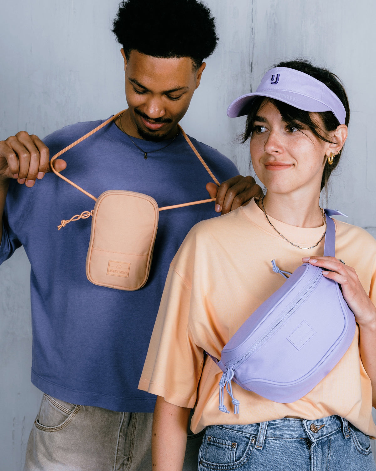 Mann trägt Umhängetasche 'Josh' in Pfirsichfarbe über blauem Shirt, Frau trägt lila Bauchtasche und Stirnband mit Johnny Urban Logo
