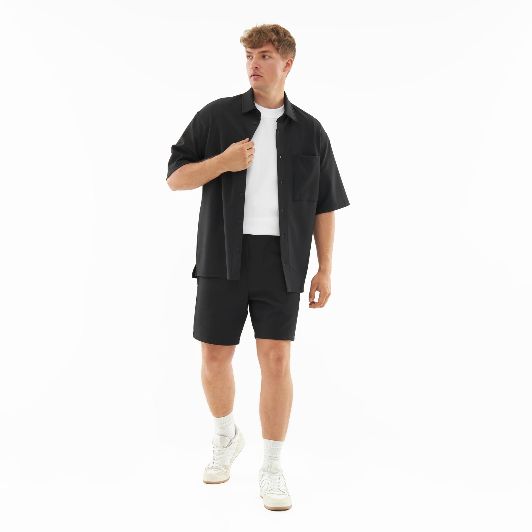 Mann trägt schwarzes, locker sitzendes Hemd und Shorts aus dem Set 'Mason', kombiniert mit weißem T-Shirt und Sportschuhen