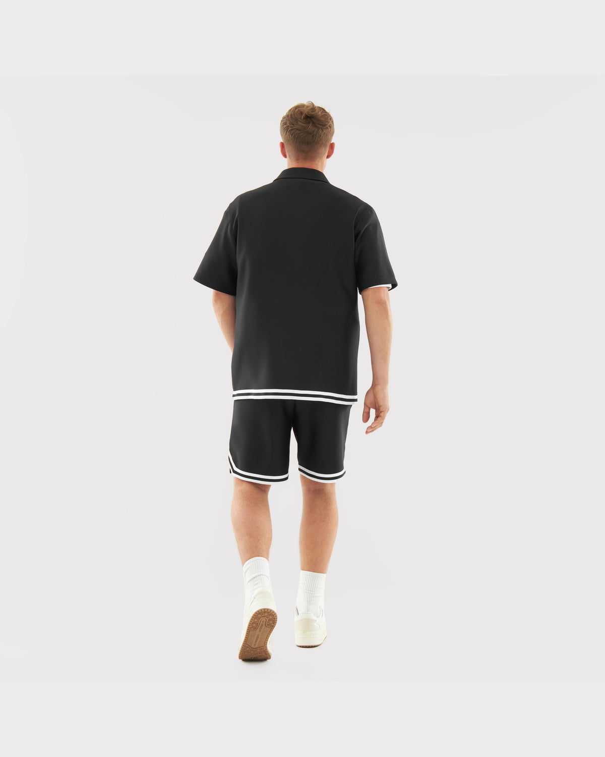 Mann von hinten in lockerem schwarzem Hemd und Shorts mit weißen Streifen am Saum, weiße Socken und Schuhe