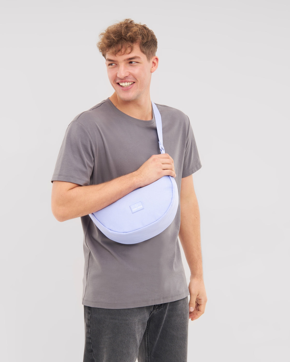 Mann trägt die lilafarbene Half Moon Bag 'Ally Medium' über der Schulter mit grauem T-Shirt und dunkler Hose