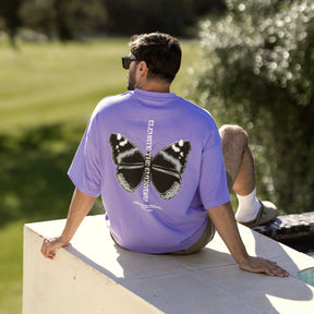 Mann sitzt draußen, trägt lila Oversize-T-Shirt 'Sammy Chrome Butterfly' mit großem schwarzem Schmetterlingsdruck auf dem Rücken