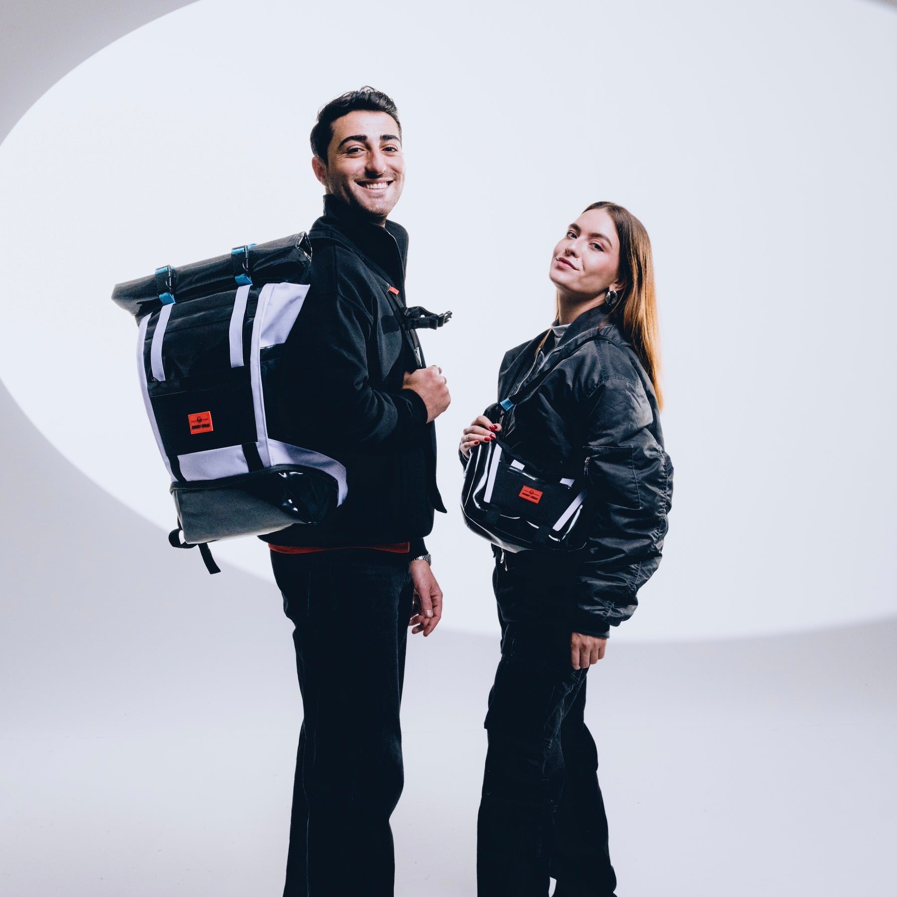 Mann mit schwarzem Rolltop Rucksack 'Ashton' und Frau mit passender Umhängetasche, beide in schwarzen Outfits