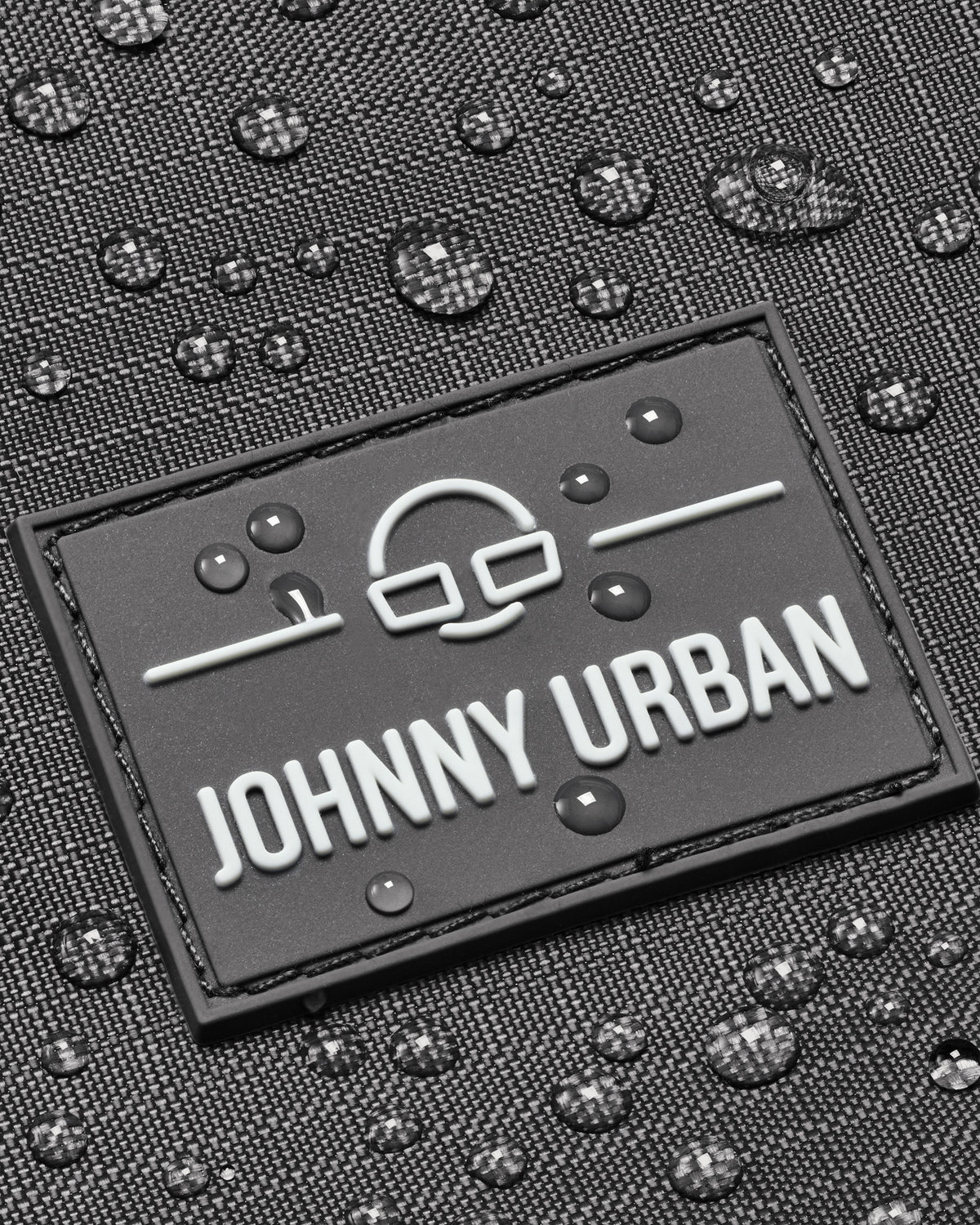 Nahaufnahme des Johnny Urban Logos auf grauem, wasserabweisendem Harper Rucksack mit Wasserperlen