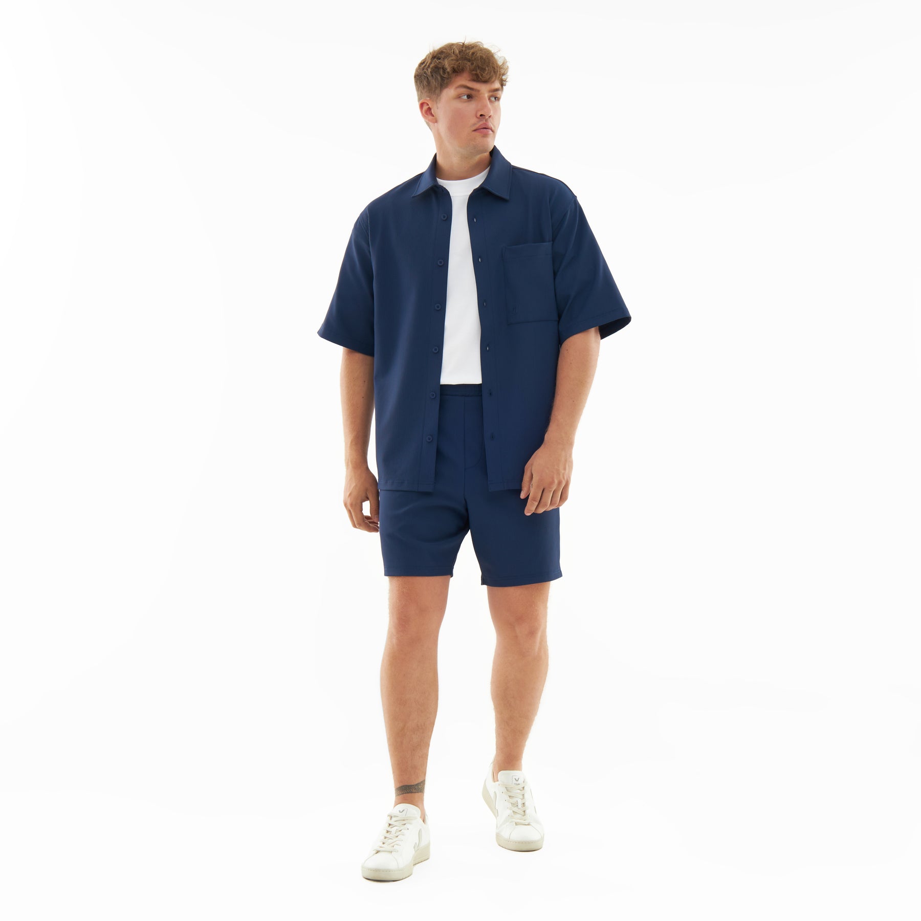 Männliches Modell trägt marineblaue Shorts 'Mason' und passendes Hemd, kombiniert mit weißem T-Shirt und weißen Sneakern