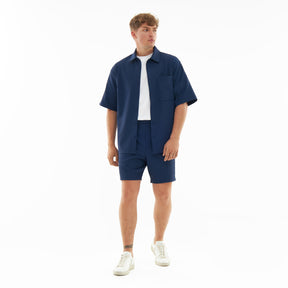 Männliches Modell trägt marineblaue Shorts 'Mason' und passendes Hemd, kombiniert mit weißem T-Shirt und weißen Sneakern