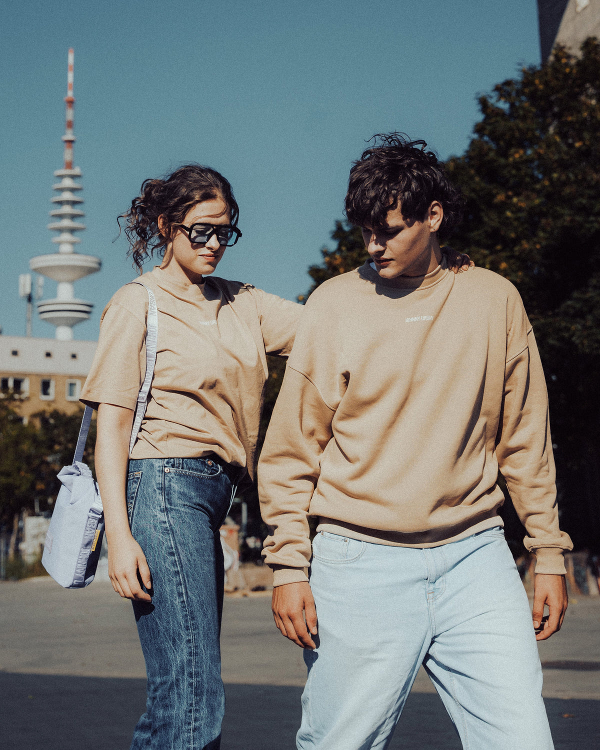 Frau in beige T-Shirt 'Sammy Johnny Urban' mit Jeans und Sonnenbrille, Mann in beige Pullover und hellen Jeans draußen in Stadtumgebung