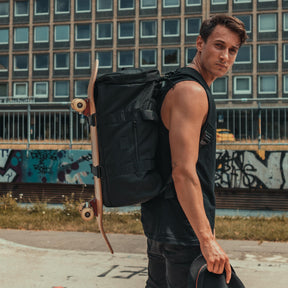 Mann mit schwarzem Duffle Bag 'Colin' und Skateboard an Rücken, hält schwarzen Hut, urbaner Hintergrund