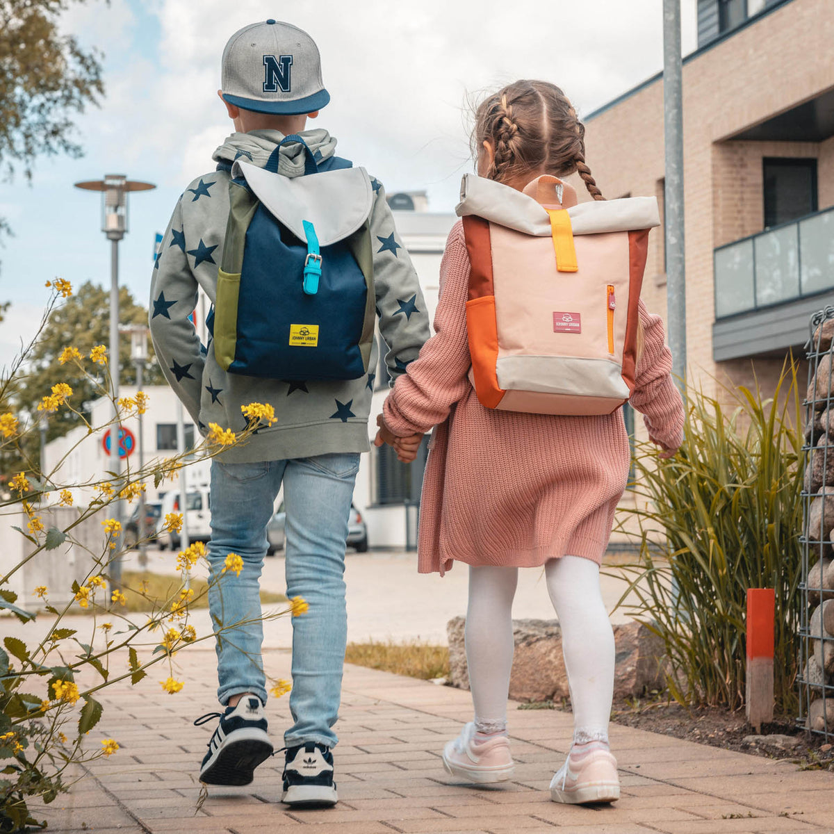 Junge mit blau-grünem und Mädchen mit rosa-orangefarbenem Kinderrucksack 'Liam' gehen Hand in Hand auf einem Gehweg