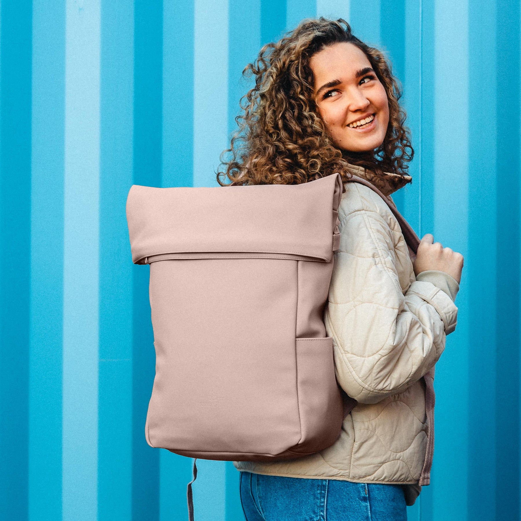 Frau mit lockigem Haar trägt rosa Johnny Urban Rolltop Rucksack Henry vor blau gestreiftem Hintergrund