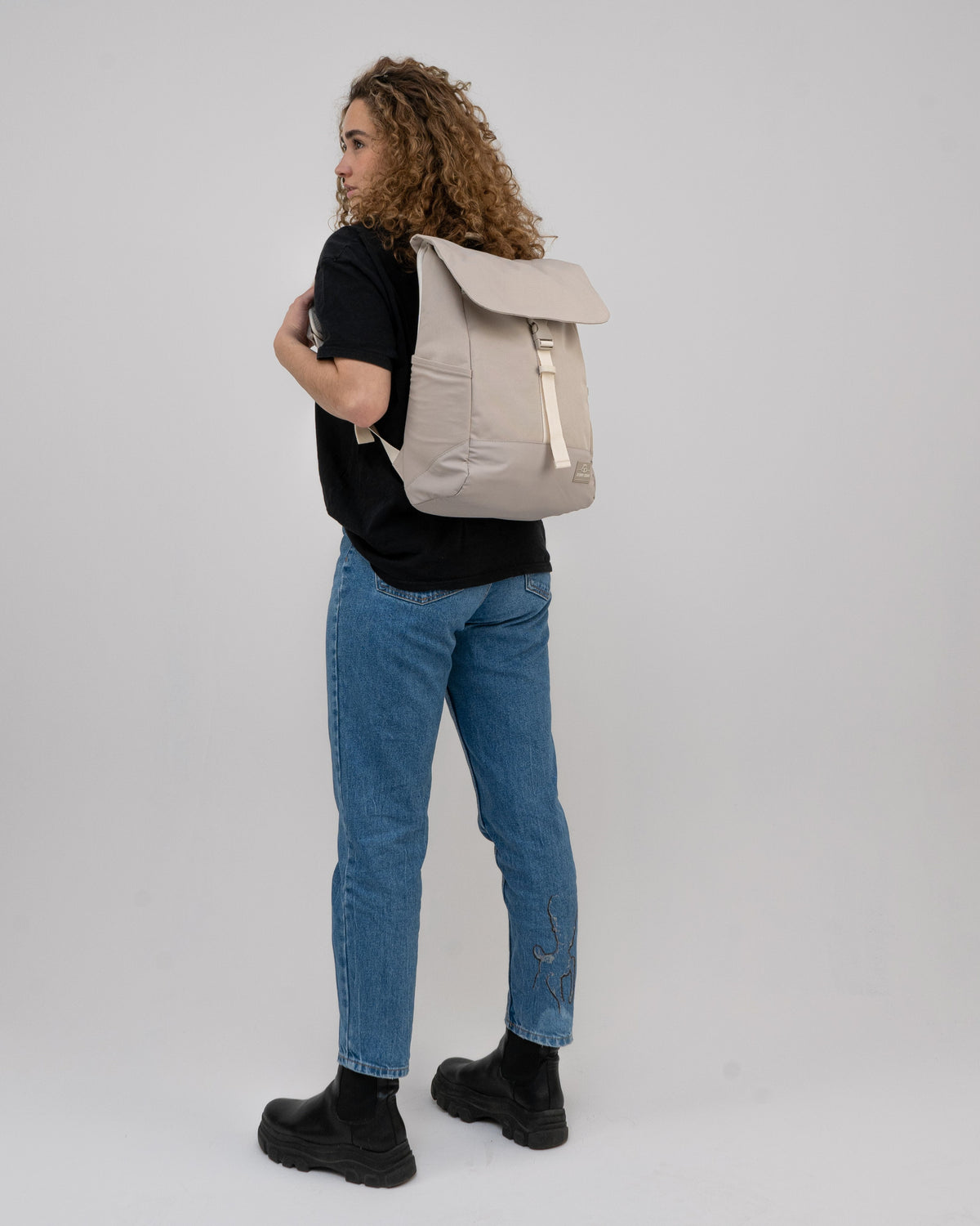 Frau mit beigem Rucksack 'Mika', blauer Jeans mit Stickerei und schwarzem T-Shirt von hinten