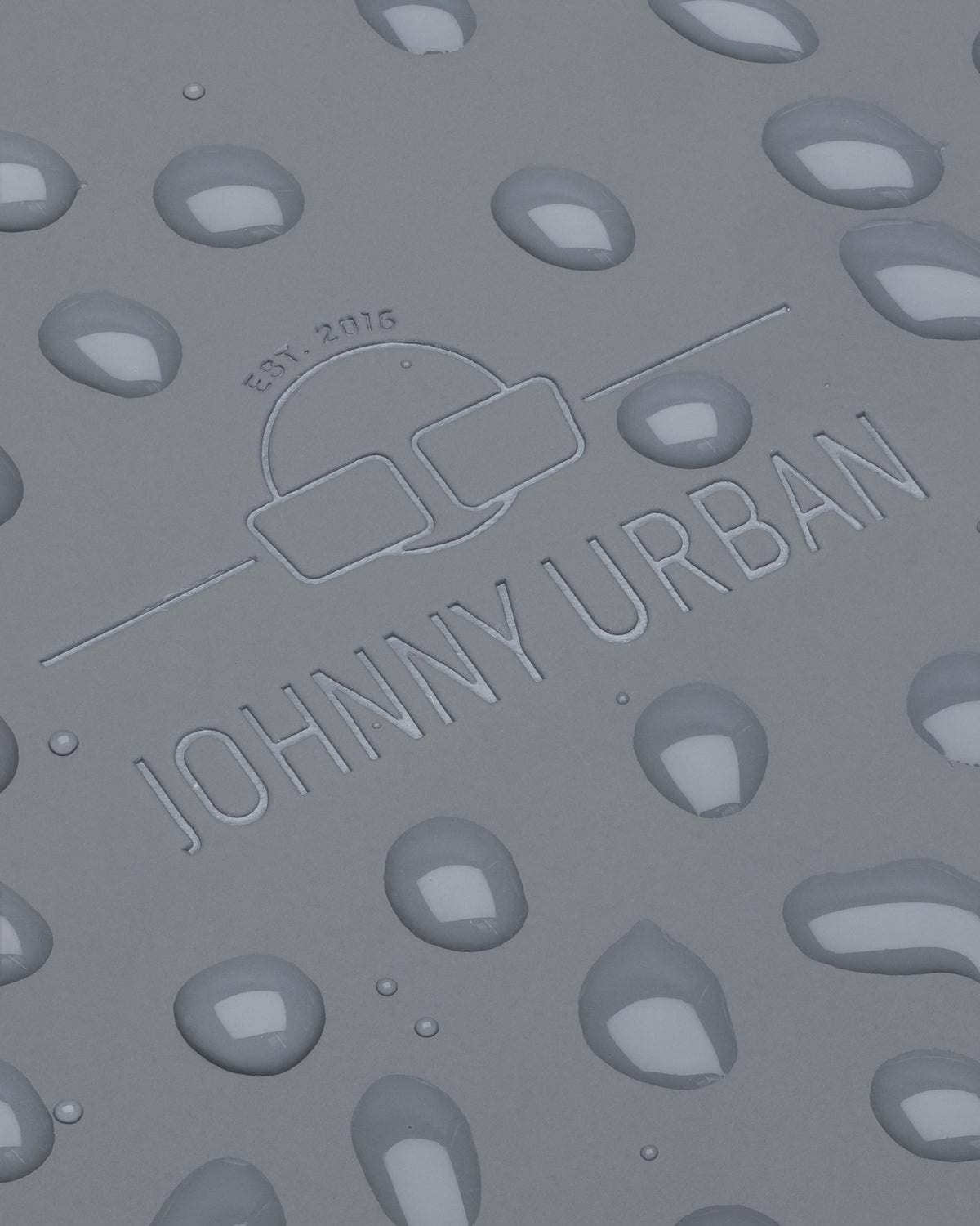 Geprägtes Johnny Urban Logo mit Sonnenbrillensymbol und Text 'Est. 2016' auf grauem Material