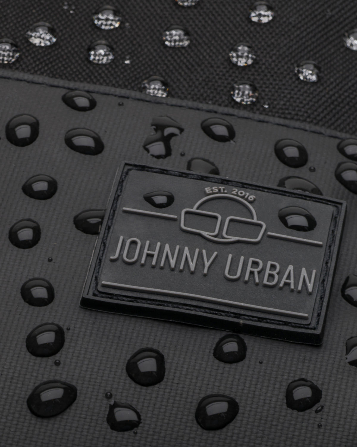 Nahaufnahme des wasserdichten schwarzen Rucksacks Mika von Johnny Urban mit Wassertropfen auf der Oberfläche und Markenlogo